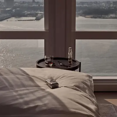 Sunlit bedroom with waterfront view, rumpled white bedding, and a bedside table with drinks and keys.
