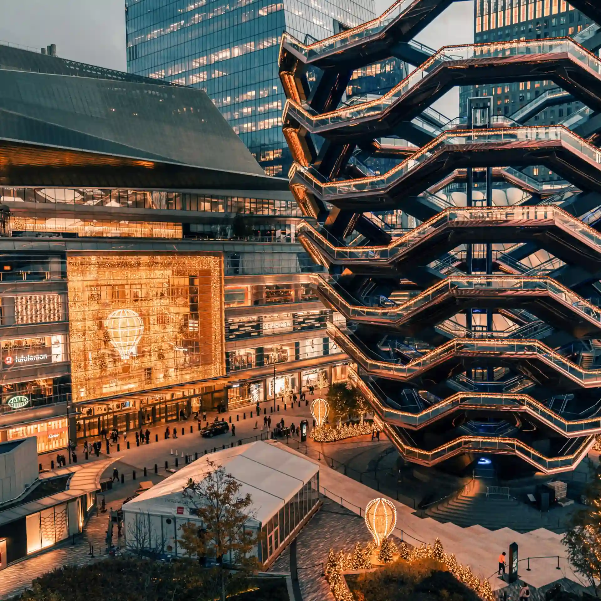 The Vessel at Hudson Yards illuminated with golden lights, surrounded by festive decorations and bustling crowd