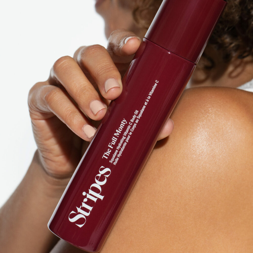 Person holding maroon bottle of body oil labeled “Stripes THE FULL MONTY” near shoulder.
