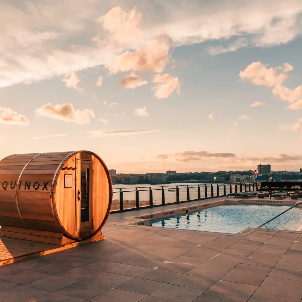 Rooftop pool at Equinox Hotel New York at sunset, featuring a wooden barrel sauna, sleek lounge chairs, glass railing, and breathtaking views of the city skyline over the river, blending luxury, wellness, and urban elegance.