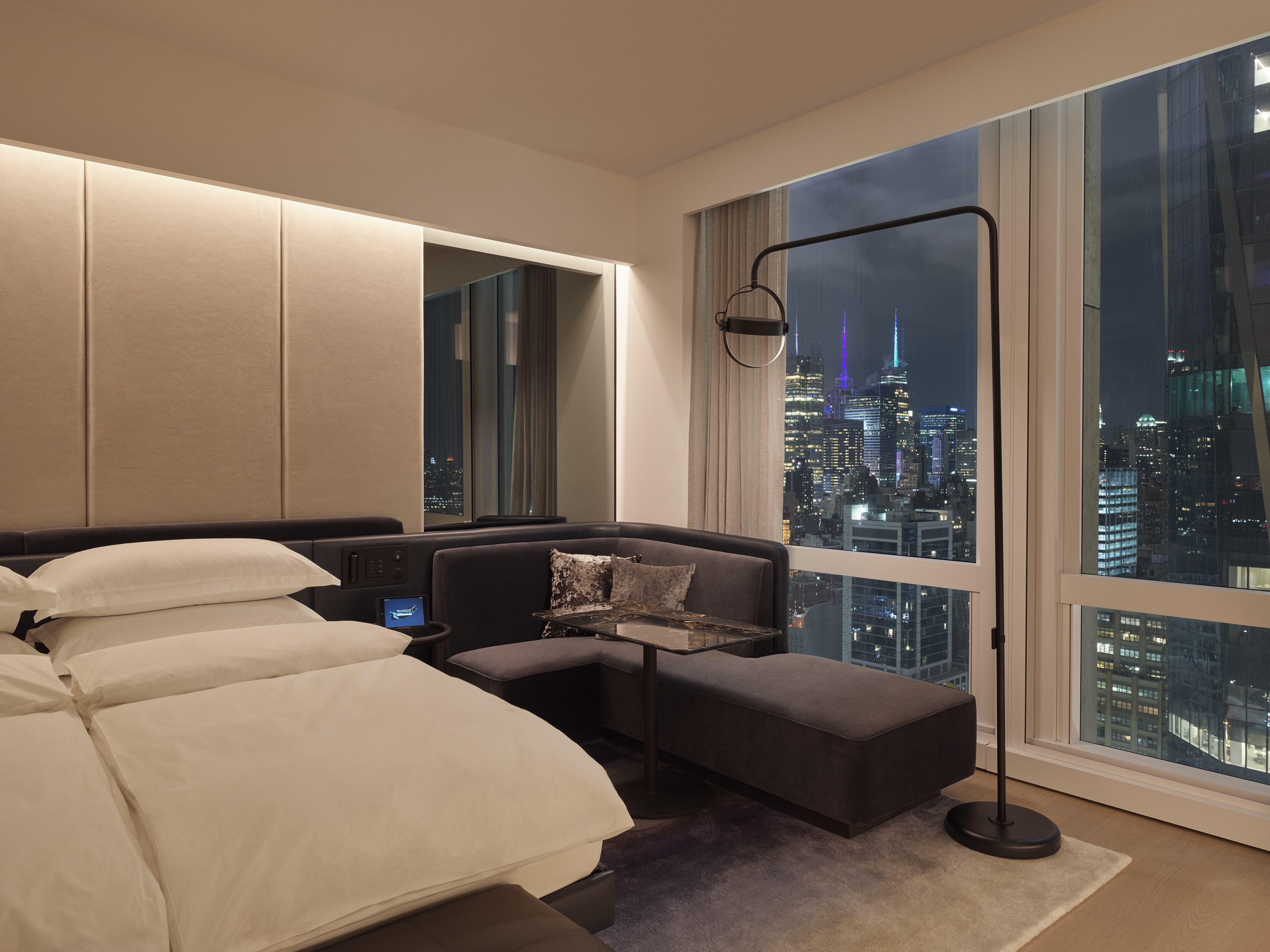 A city-view king room after dark, the bed and a dark upholstered sofa lit by a single arc lamp, with the lit towers of Manhattan filling the corner windows.