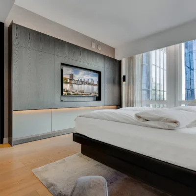 Upscale bedroom with white linens, floor-to-ceiling windows, city view, and mounted TV above lit cabinet.