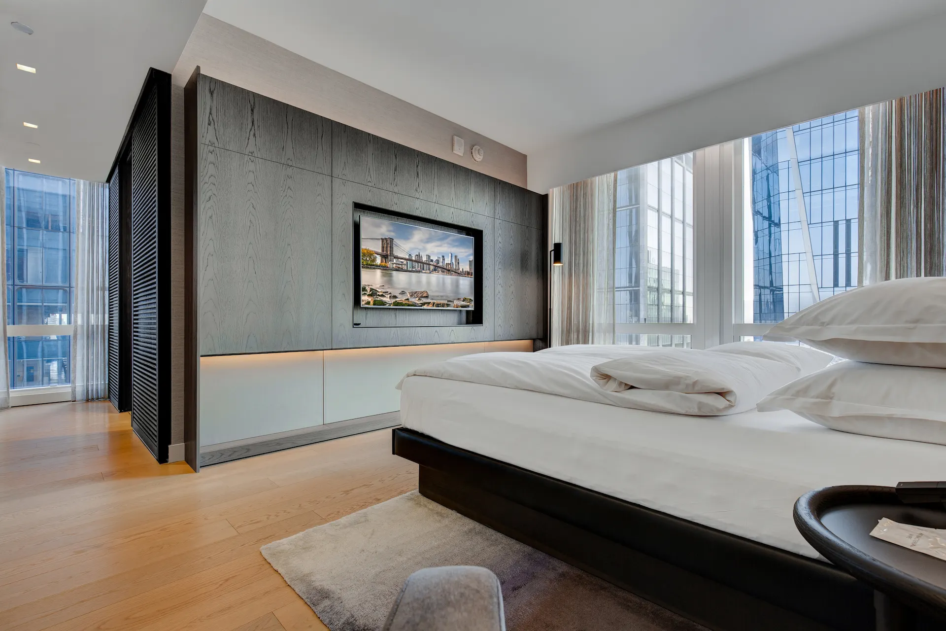 Upscale bedroom with white linens, floor-to-ceiling windows, city view, and mounted TV above lit cabinet.