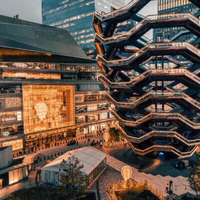 The Vessel at Hudson Yards lit with warm lights, surrounded by skyscrapers, people, and festive balloon displays.