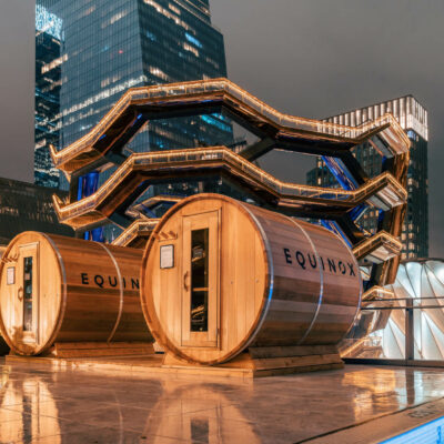 Rooftop terrace with wooden sauna pods labeled "EQUINOX" and illuminated Vessel structure in Hudson Yards at night.