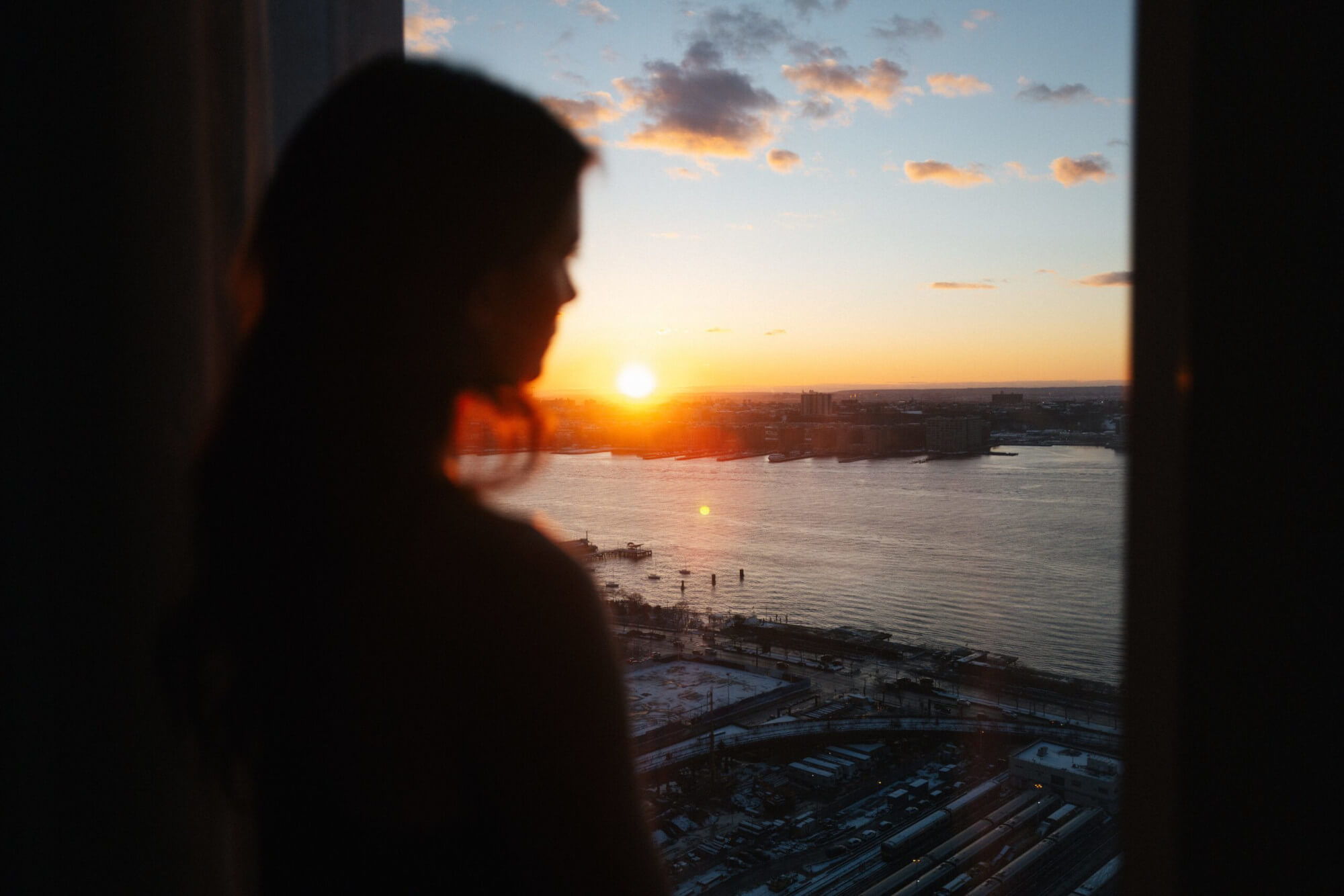 Person stands by window watching sunset over water, silhouetted against warm orange sky and distant cityscape.