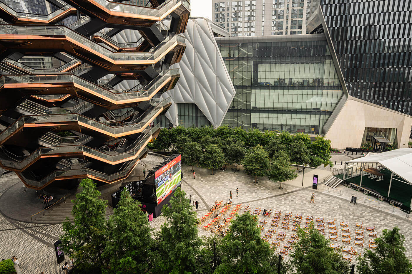 Urban plaza featuring the Vessel and The Shed in Hudson Yards, with orange chairs and digital screen set for public event.