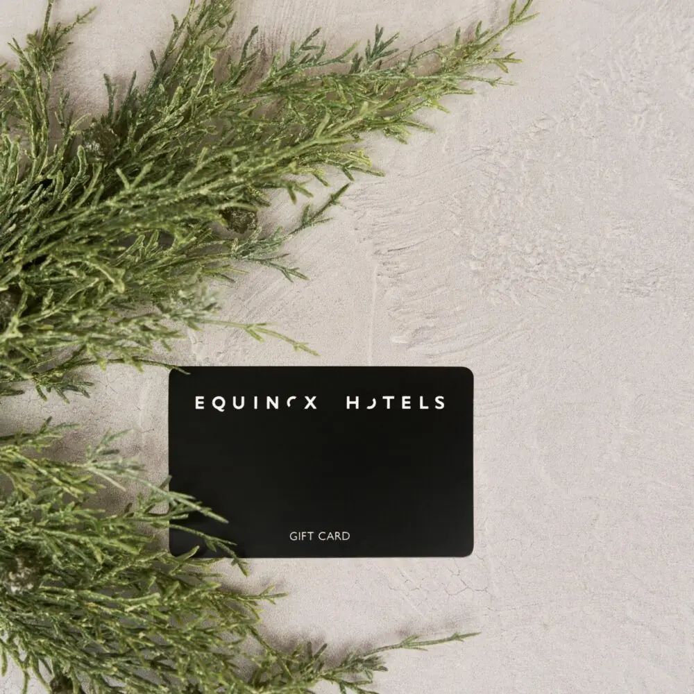 Black gift card labeled "EQUINOX HOTELS" with stylized text, placed on textured surface and surrounded by green foliage.