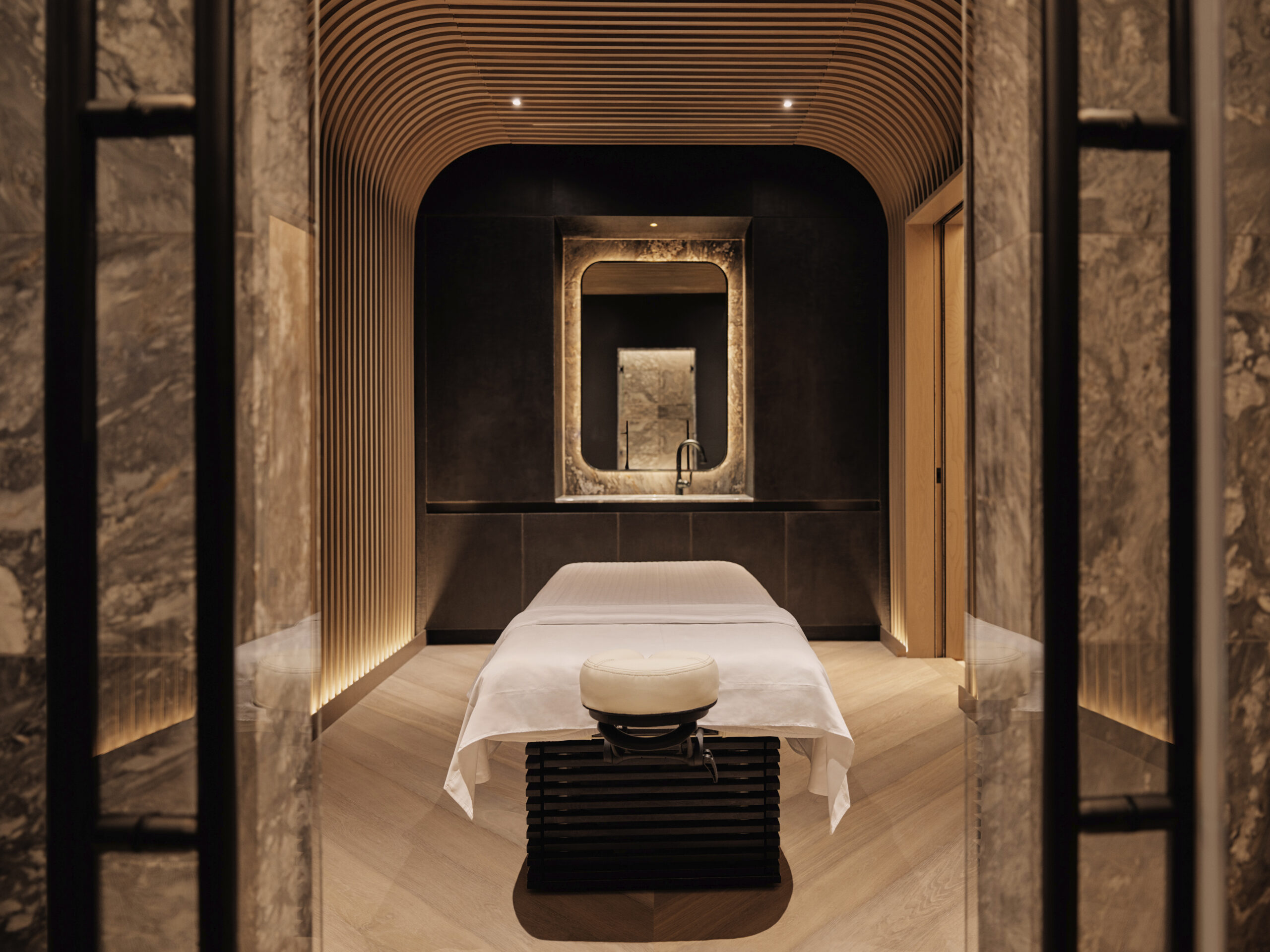 A spa treatment room framed by fluted stone piers, a single dressed massage table under a curved slatted ceiling with a backlit niche on the far wall.