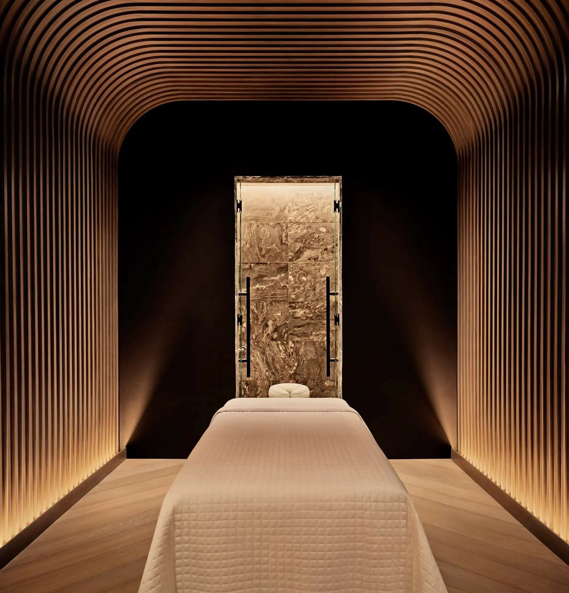 Massage therapy room with curved wood architecture, ambient lighting, and a white-sheeted table facing a textured focal wall.