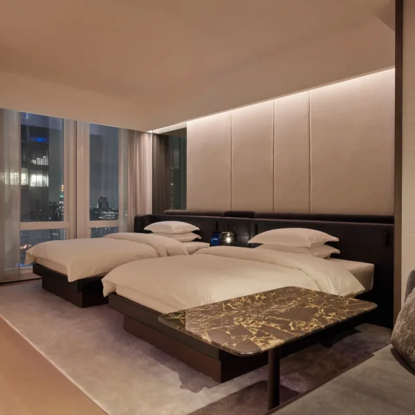 Upscale hotel room with twin beds, marble table, and nighttime city views through floor-to-ceiling windows