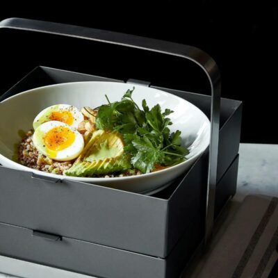 Healthy quinoa bowl with avocado, soft-boiled eggs, parsley, and stylish lunchbox on marble surface.