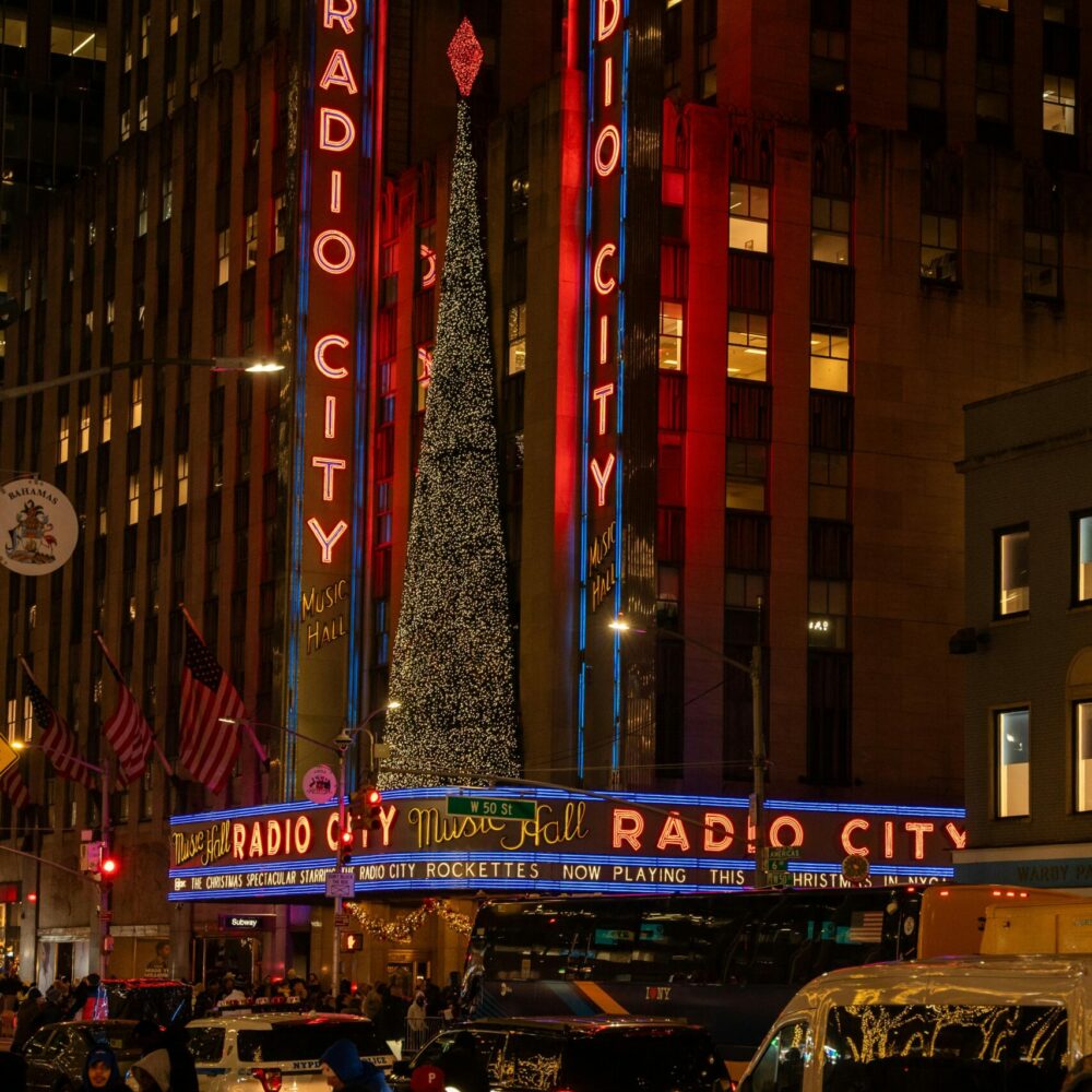 Radio City Music Hall lit up for Christmas with tree above marquee and crowd in festive New York street.
