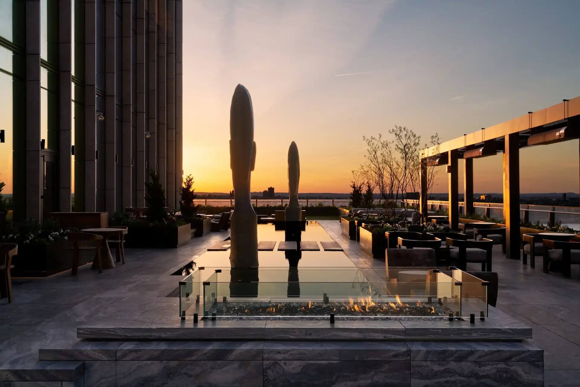 Rooftop terrace at sunset with fire pit, sculptures, and water views, evoking artful serenity and elevated design