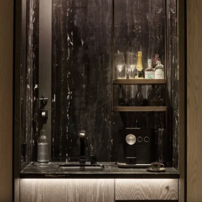 Minibar at Equinox Hotel with dark marble backsplash, black sink and faucet, coffee machine and bottles on wooden shelves.
