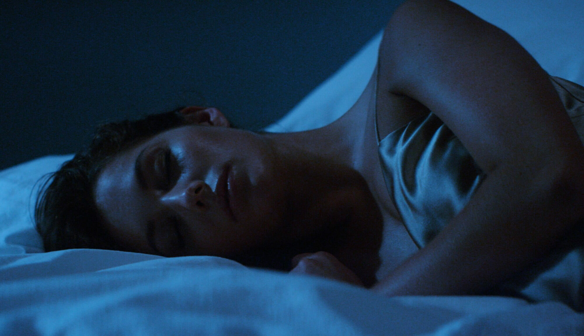 Person resting in white bed at Equinox Hotel, wearing satin sleepwear, bathed in cool blue light for a calm, restorative mood