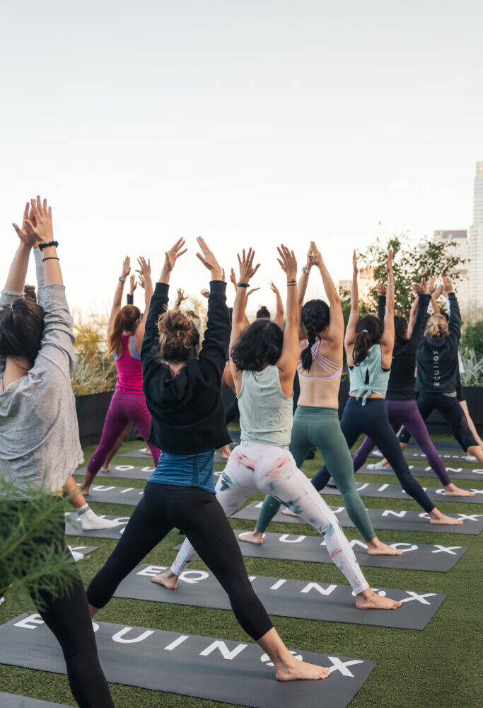 Outdoor rooftop yoga class with participants in athletic wear, arms raised, city buildings and plants in background.