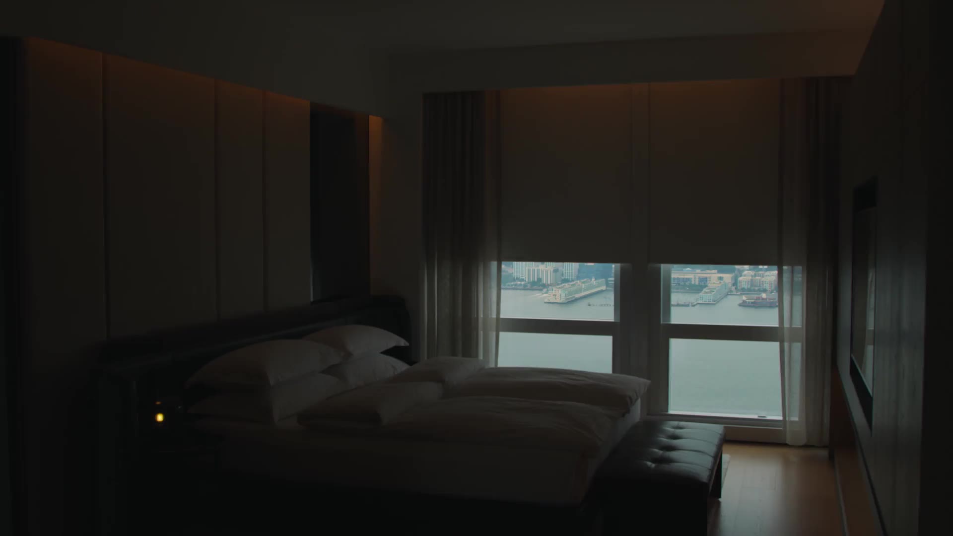 Modern bedroom with white bedding, ambient lighting, and harbor view through floor-to-ceiling windows.