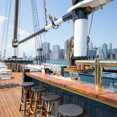 Sailing ship deck converted into a bar and dining area with stools, tables, and city skyline in the background.