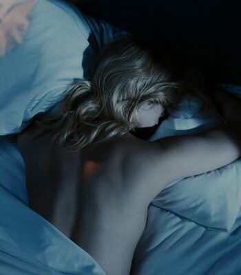 Person lying face down on white bed at Equinox Hotel, back exposed, head turned to side, in dim light creating a calm.