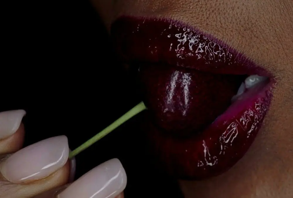 Red lips holding a cherry between the teeth, set against a neutral background; close-up detail emphasizing texture, contrast, and sensuality