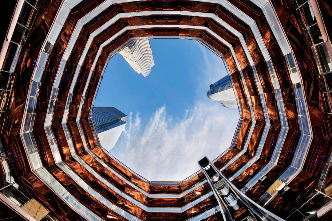 Upward view from inside the Vessel at Hudson Yards showing copper staircases and framed sky with skyscrapers
