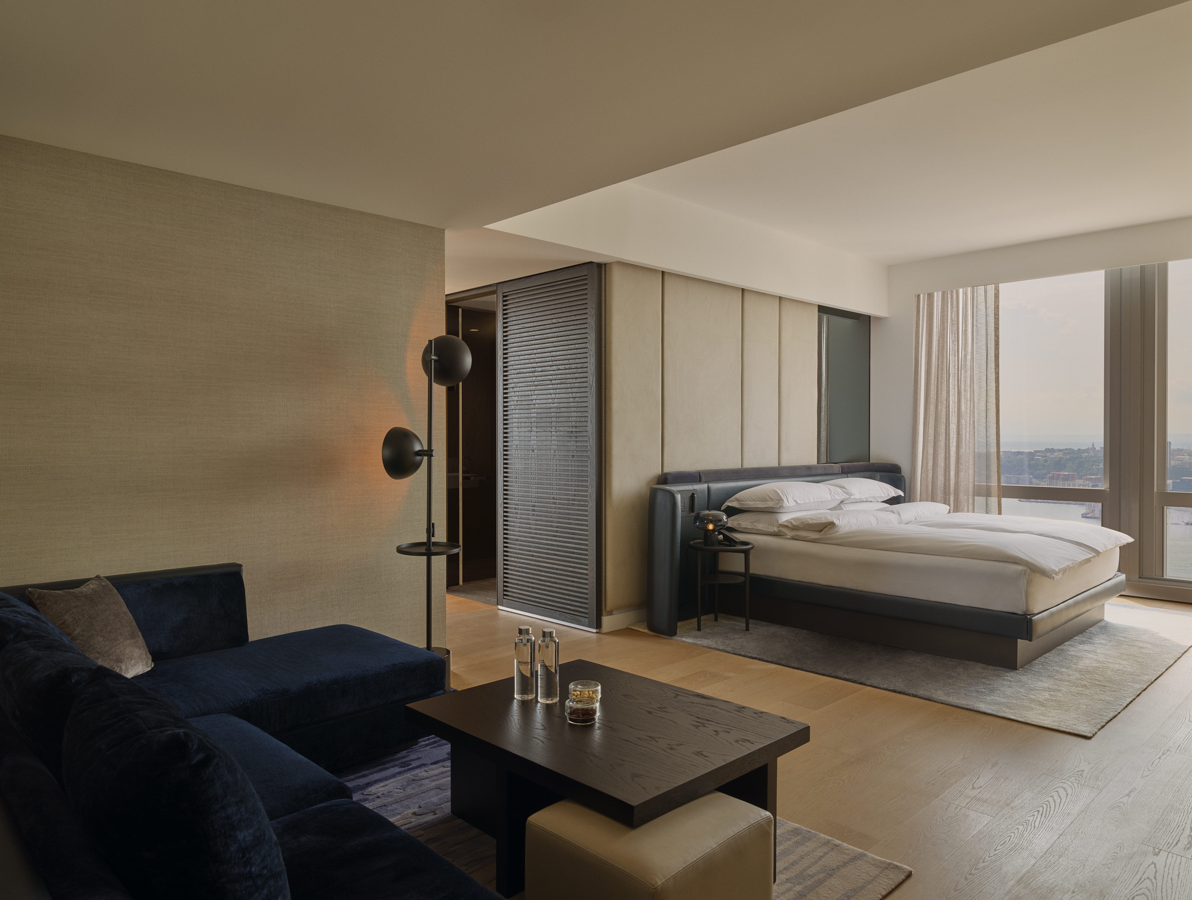 A junior suite with a dark sofa and low table in the foreground and the bed set behind a partial wall, daylight from windows onto the river.