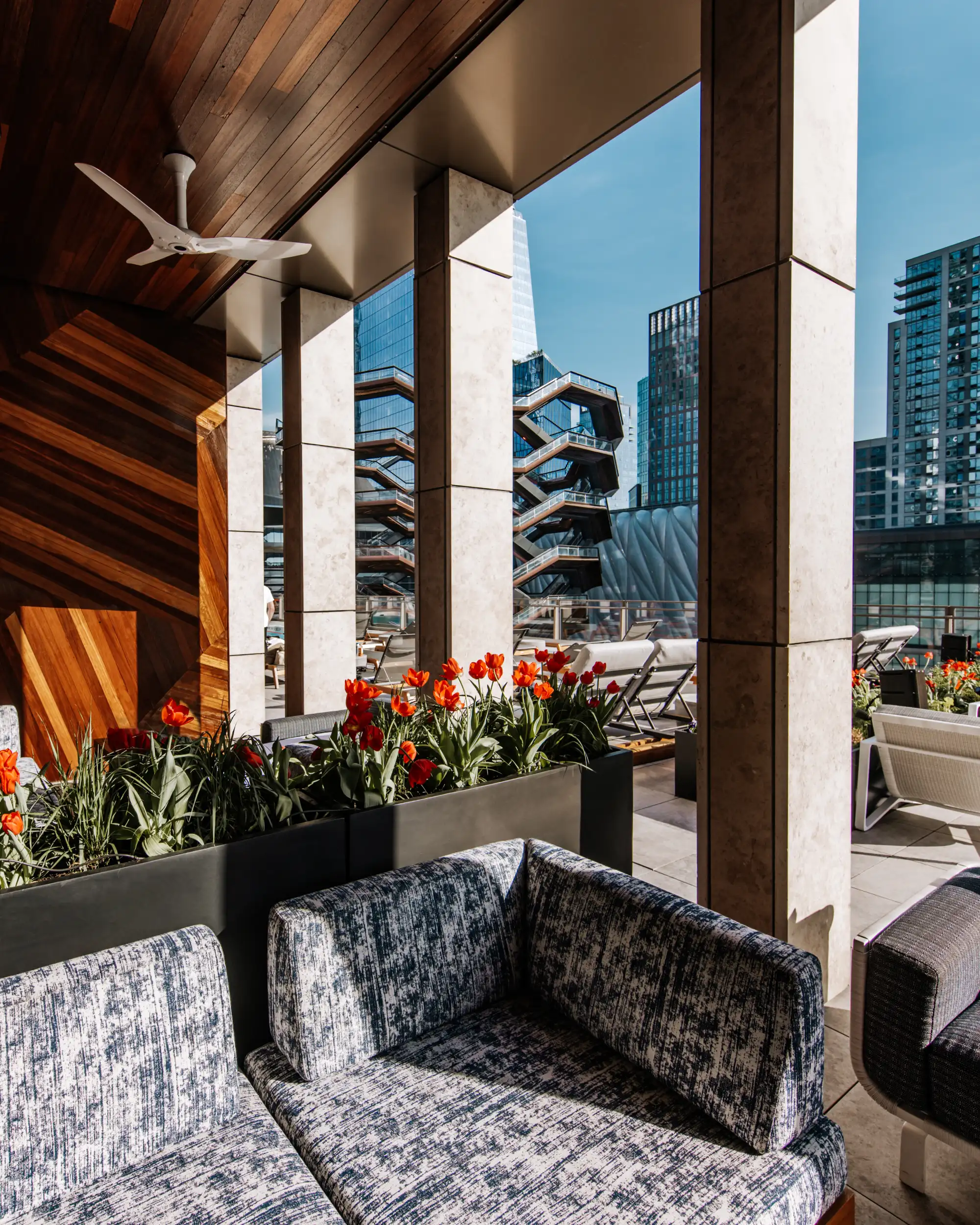 A terrace seating area under a timber soffit, patterned sofas beside planters of red tulips, looking out between concrete columns to the Vessel and the towers of Hudson Yards.