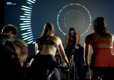 Group spin class in dark room with large screen showing pulsating light patterns, instructor leading high-energy session