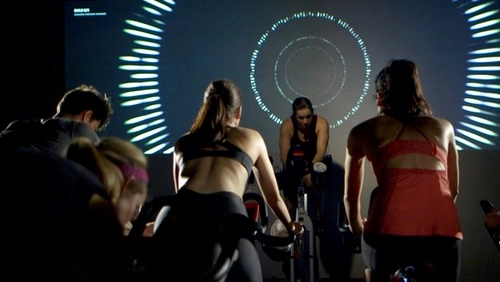 Group spin class in dark room with large screen showing pulsating light patterns, instructor leading high-energy session