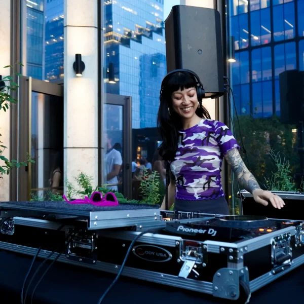 DJ in purple camo shirt performing outdoors with headphones and tattoos, framed by glass buildings and greenery in urban setting