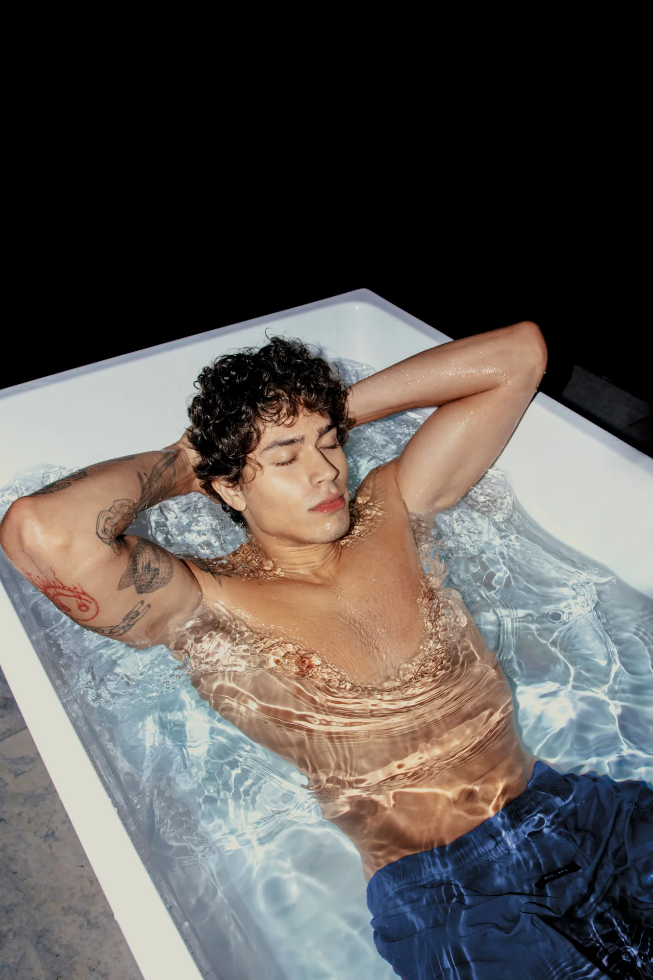 Person with tattoos relaxing in white tub filled with water, light reflections on skin in dark setting highlighting calm and contrast