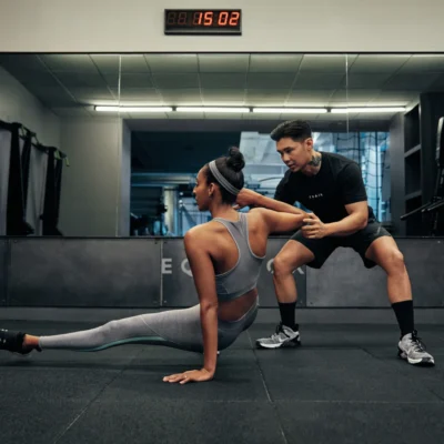 Woman doing resistance band floor exercise with male trainer assisting in well-lit gym