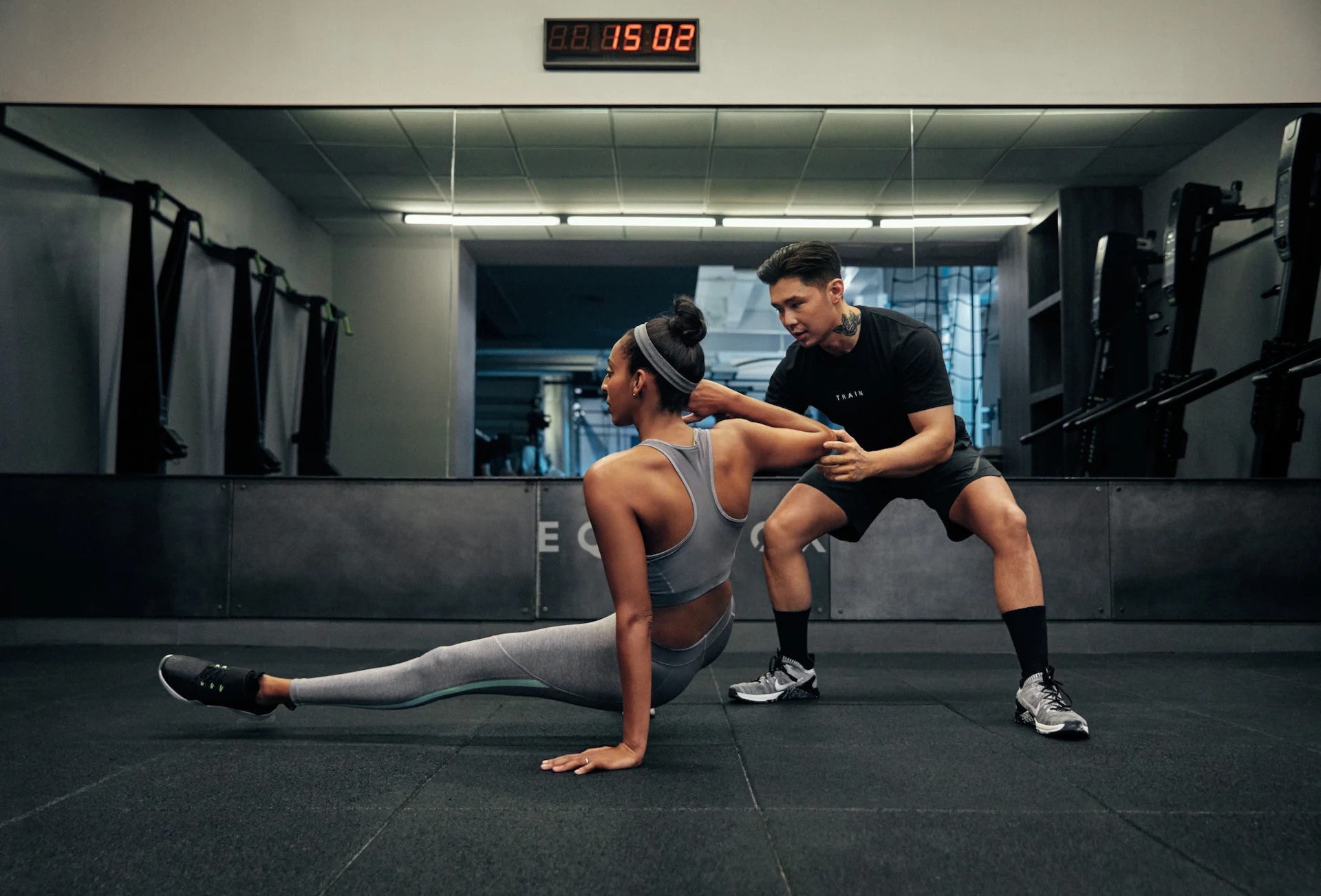Woman doing resistance band floor exercise with male trainer assisting in well-lit gym