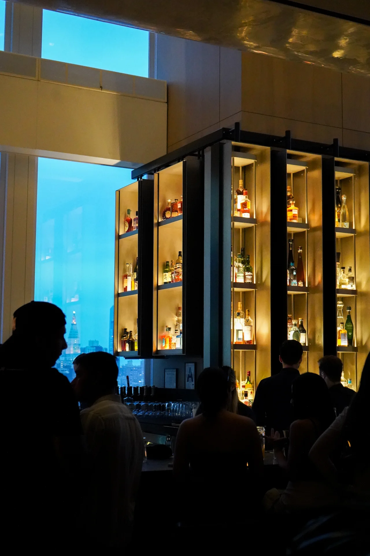 Evening bar scene with backlit liquor shelves, city views, and silhouetted guests