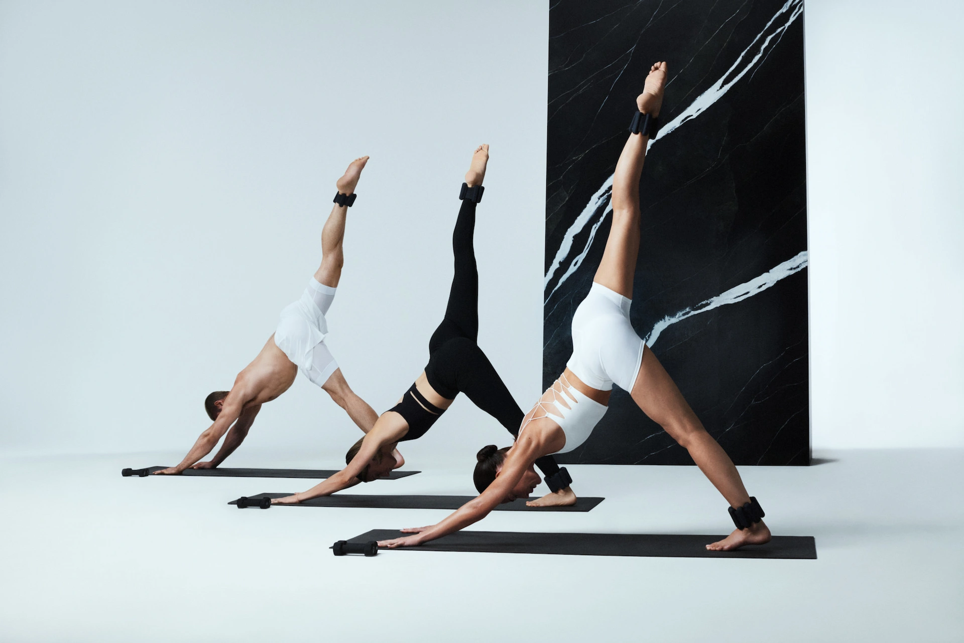 Three individuals in three-legged downward dog pose with ankle weights, set against minimalist studio backdrop, evoking strength, balance, and elevated movement