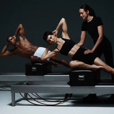 Three people doing Pilates on reformer machines, with instructor assisting posture in minimal studio with dark background