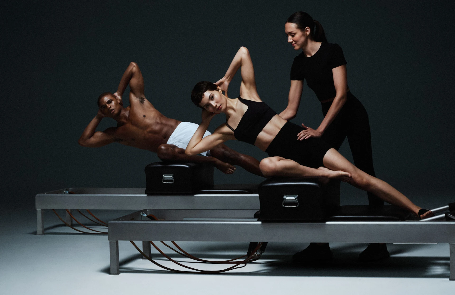 Three people doing Pilates on reformer machines, with instructor assisting posture in minimal studio with dark background