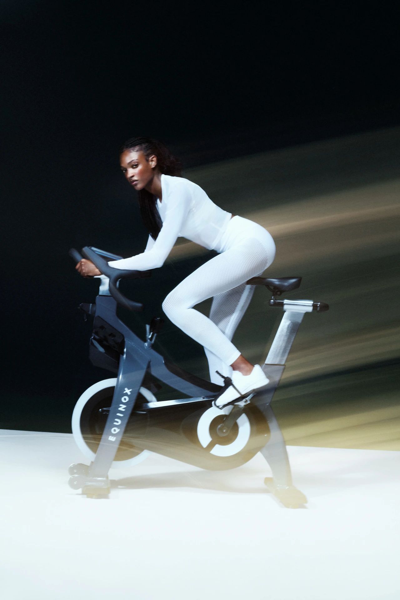 Woman riding Equinox cycling bike in white athletic wear, framed by motion light and focused intensity