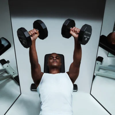 Person doing dumbbell bench press on gym bench, wearing white sleeveless shirt, mirrors in background.