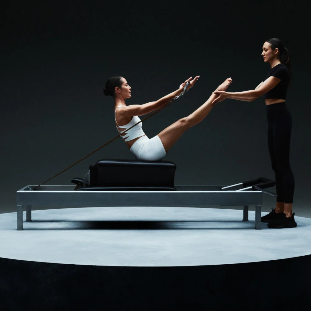 Person performing V-sit Pilates move on reformer with instructor supporting feet, set on circular platform against dark background