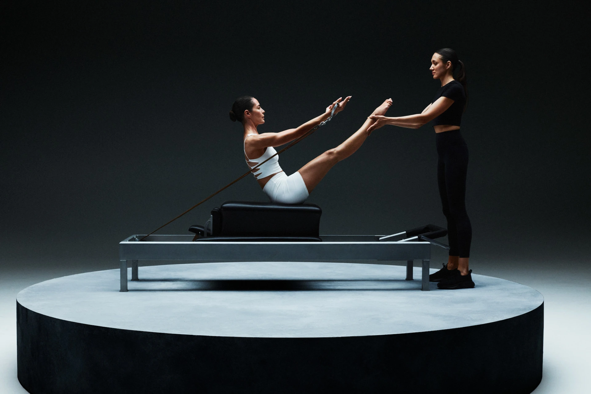Person performing V-sit Pilates move on reformer with instructor supporting feet, set on circular platform against dark background