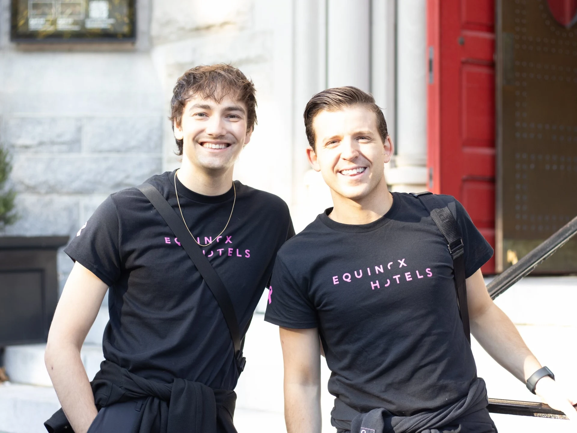 Two Equinox Hotels team members smiling outdoors in branded shirts, standing by red door and stone facade in urban setting.