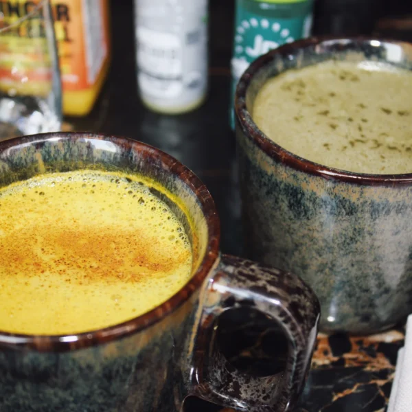 Two ceramic mugs on marbled surface with yellow turmeric drink and green herbal beverage, bottles in background.