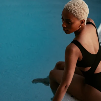 Person in black swimsuit sitting poolside with feet in water, looking over shoulder at clear blue pool.