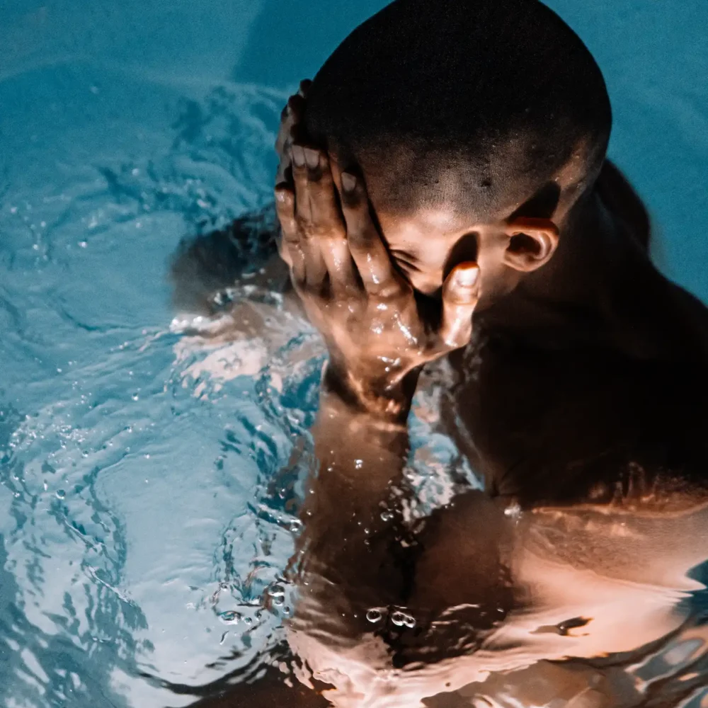 Person submerged in water with hands covering face, ripples and light highlighting muscles in dramatic, introspective setting