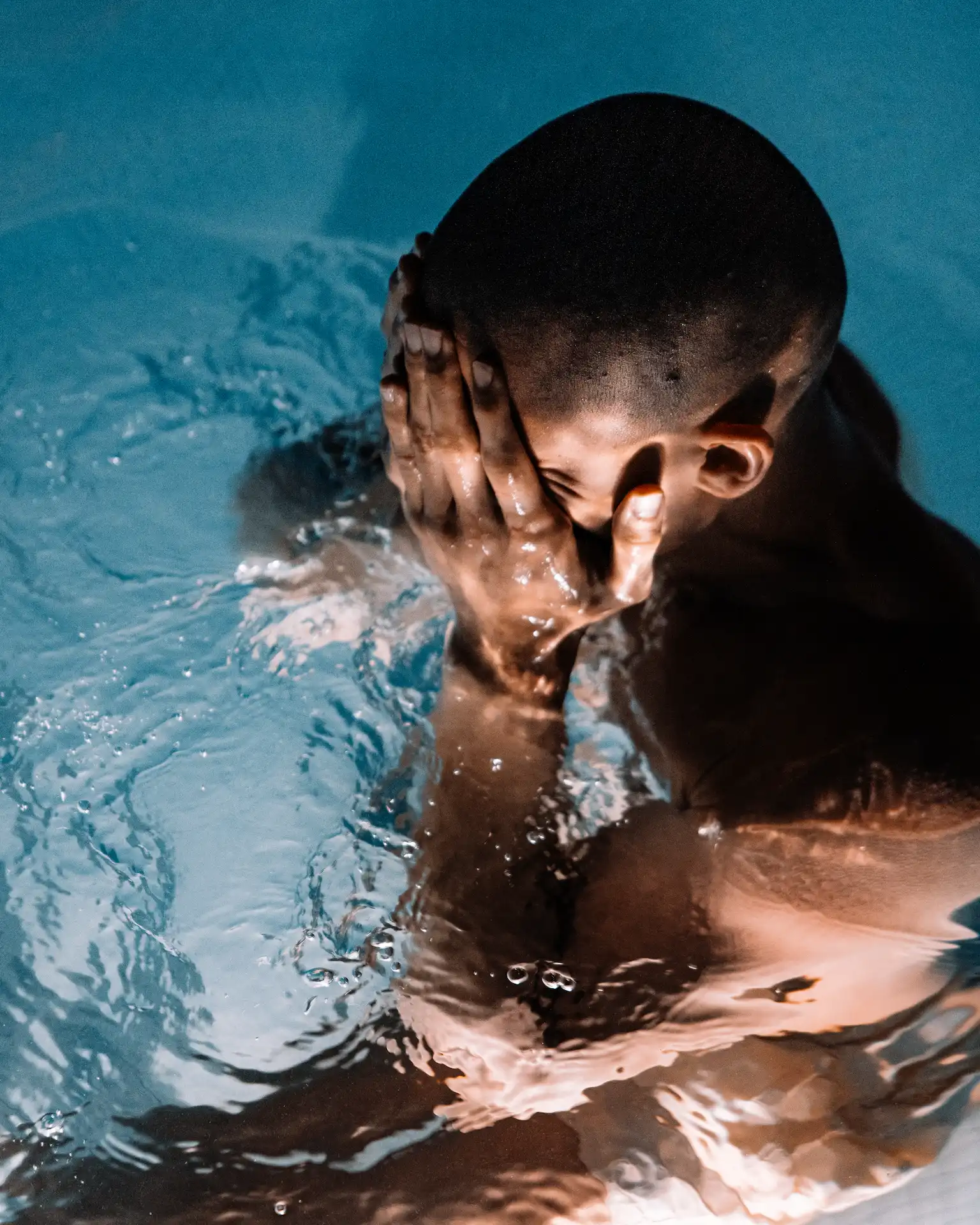 Person submerged in water with hands covering face, ripples and light highlighting muscles in dramatic, introspective setting