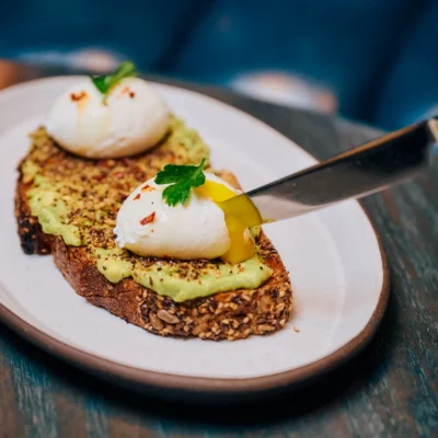 Close-up of avocado toast at Proper Hotel Weekend Brunch, featuring creamy smashed avocado topped with perfectly poached eggs, vibrant red pepper flakes, fresh parsley, and seasoned multigrain bread, served on a rustic wooden platter.