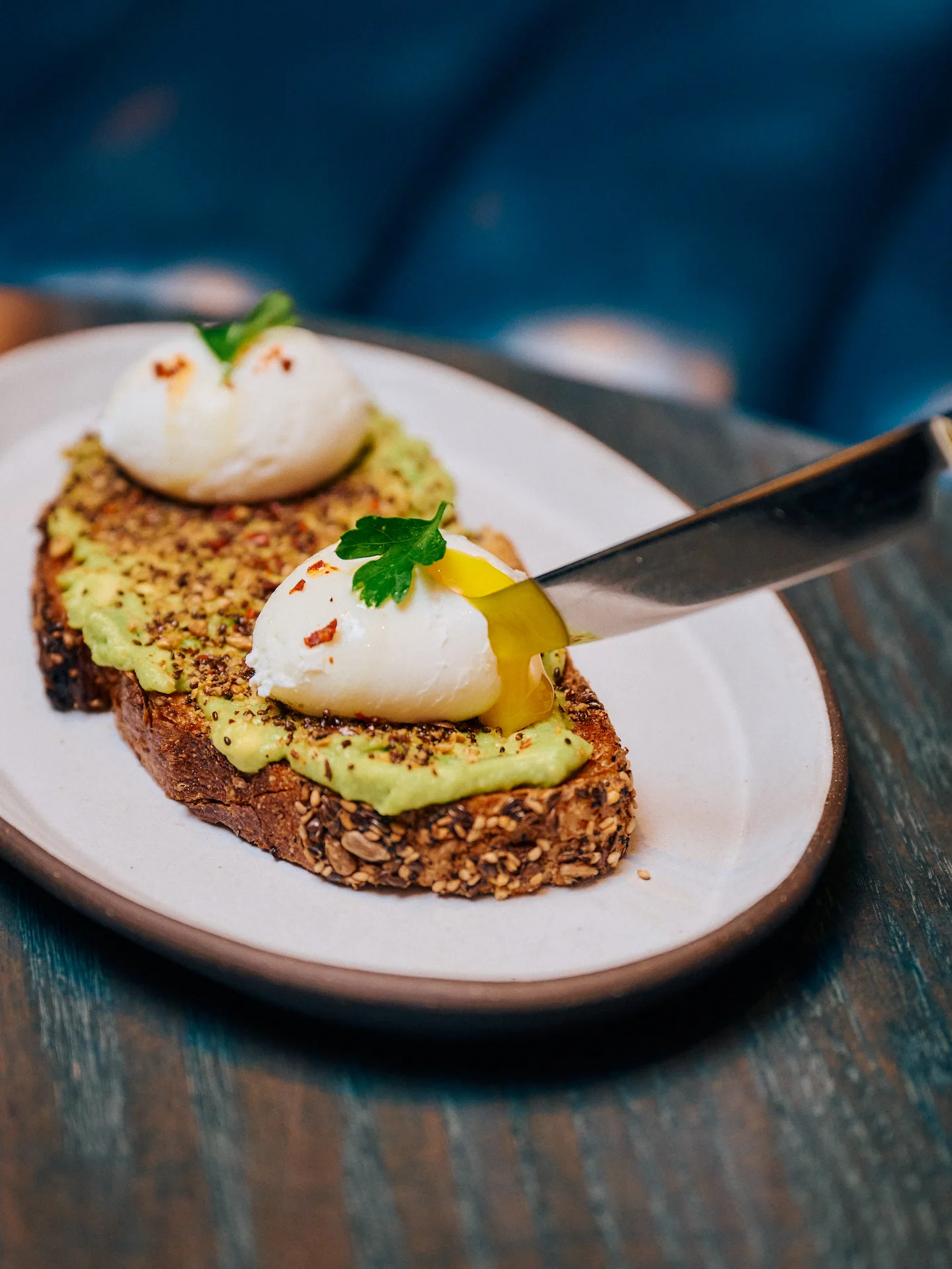 Close-up of avocado toast at Proper Hotel Weekend Brunch, featuring creamy smashed avocado topped with perfectly poached eggs, vibrant red pepper flakes, fresh parsley, and seasoned multigrain bread, served on a rustic wooden platter.