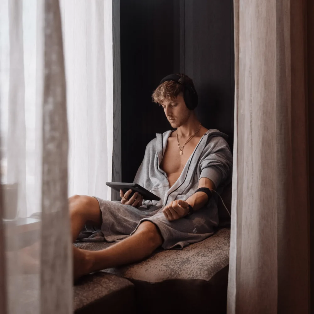 Person in grey robe on window seat with headphones, tablet, and blood pressure cuff in serene, softly lit setting.